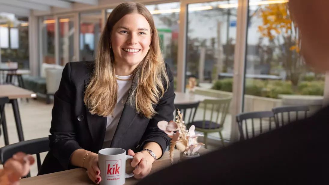 Žena sedí s úsměvem u stolu, drží v ruce šálek s logem „kik“ a vede rozhovor.