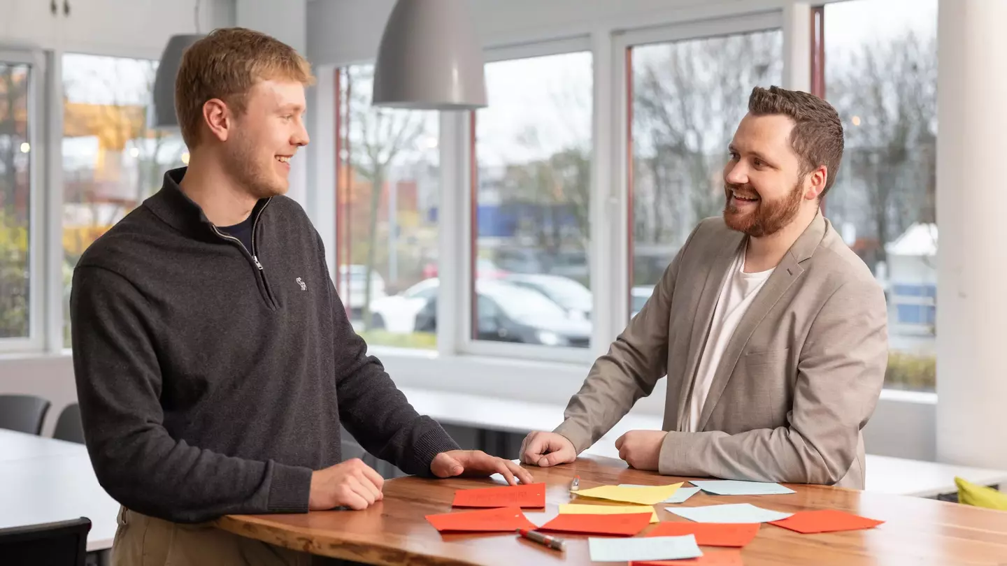 Zwei Mitarbeiter stehen lächelnd in einem modernen Büro an einem Tisch mit Post-Its