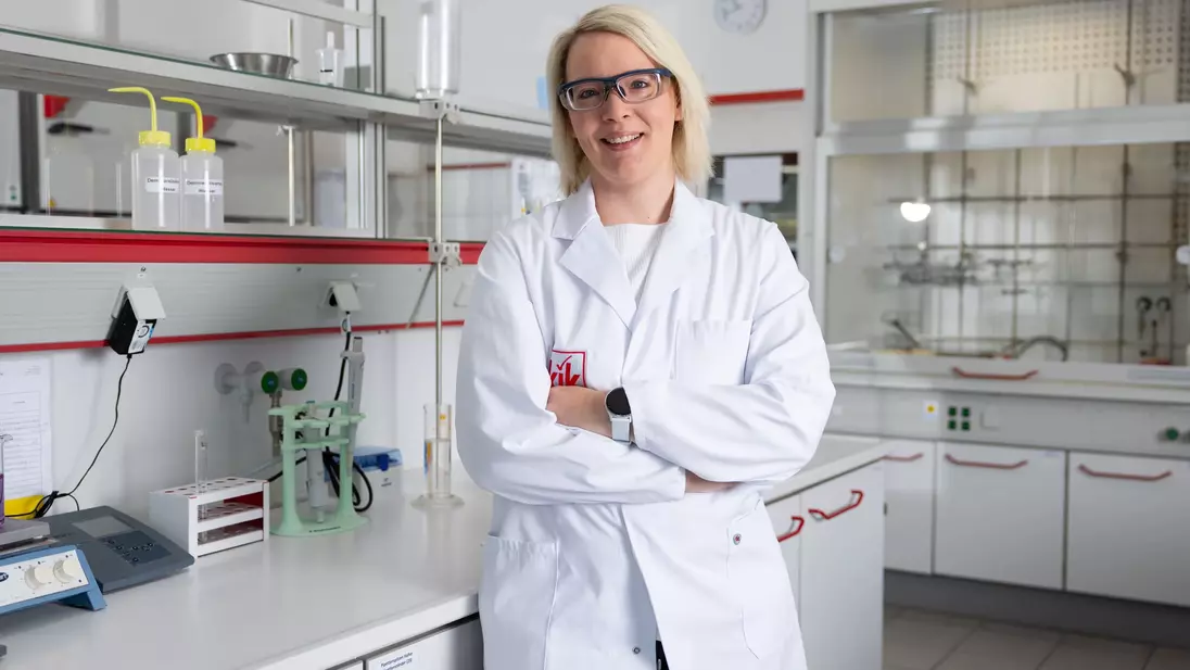 Laborantka v bílém plášti stojí se založenýma rukama v moderní laboratoři, má na sobě ochranné brýle a usmívá se.