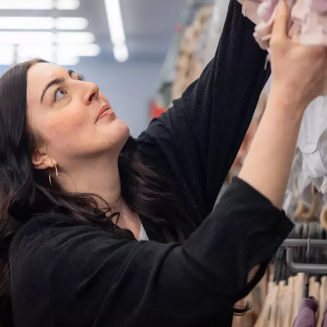 Žena s tmavými vlasy sahá nahoru do regálu a pozorně něco hledá.