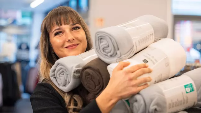 Prodavačka stojí s úsměvem v prodejně a v rukou drží spoustu zboží