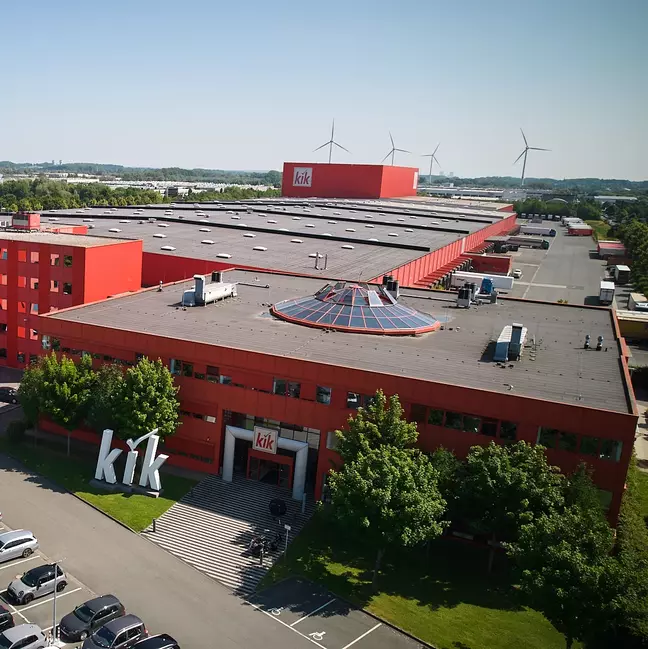 Velká červená firemní budova s logem „kik“ stojí uprostřed zeleně.