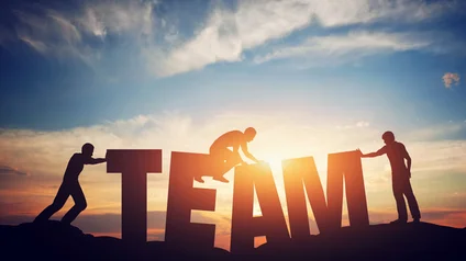 Team Es sind drei Menschen bei Sonnenuntergang zu sehen, die auf vier großen Buschstaben herumklettern, welche das Wort "TEAM" ergeben.