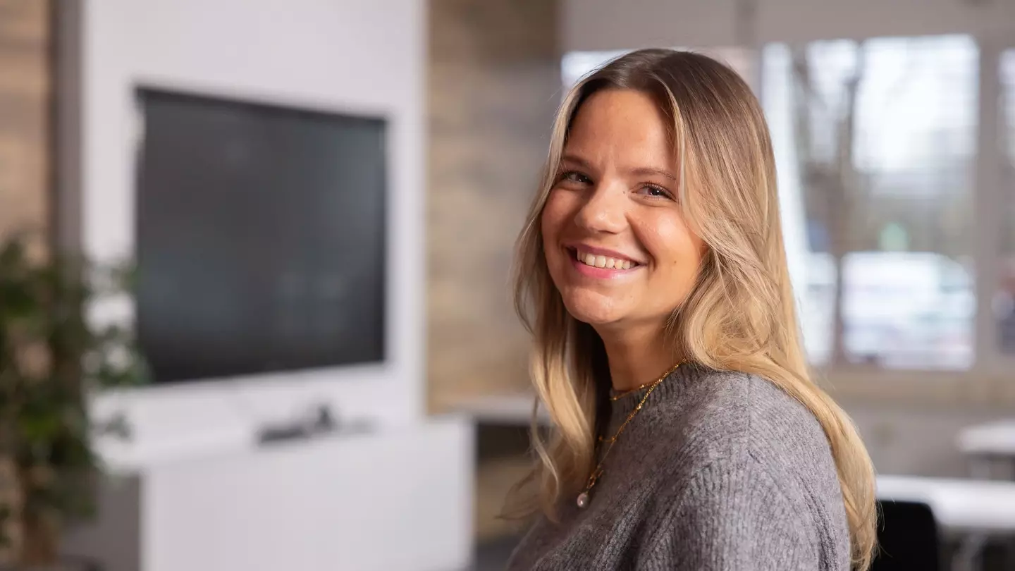 Žena se s úsměvem dívá do kamery, stojí v moderním interiéru