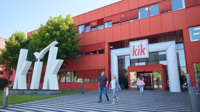 Červená budova KiK s velkým bílým logem před vchodem a několik lidí směřujících do budovy.