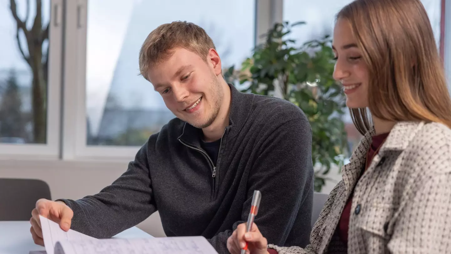 Ein Mann und eine Frau schauen in ein Notizbuch, die Frau hält einen Kugelschreiber, beide lächeln und sitzen in einem modernen Büro